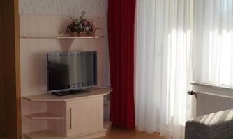 A modern living room with a TV on a light sideboard. Light curtains hang in front of the window and there is a decorative flower wreath on a shelf.