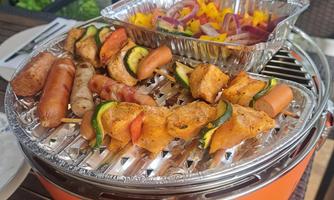 A grill with versatile dishes, including skewers, sausages, and vegetables. In the background is a bowl of colorful salad.