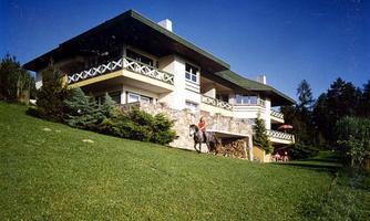 A modern house with large windows and a wide balcony. In the foreground, there is a rider on a horse on a green meadow.