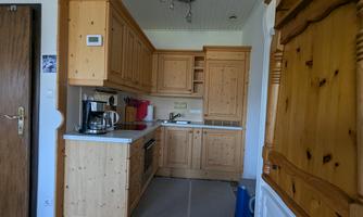 A modern kitchen with wooden furniture and white countertops. The equipment includes a stove, a coffee machine, and various kitchen cabinets.