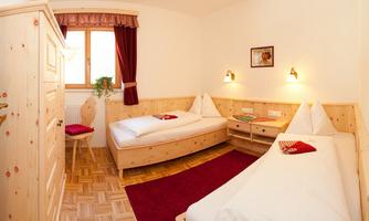 A cozy room with two single beds and wooden furniture. The room has a window with red curtains and warm light.