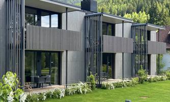 Modern residential buildings with wooden cladding and large window fronts. Surrounded by green landscape and well-maintained lawn.