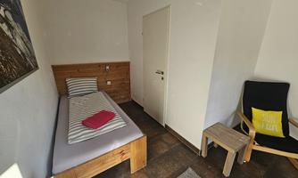 A simple room with a single bed, a chair, and a small table. The walls are light, and there is a touch of wooden decor.