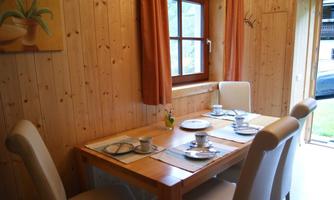 A cozy dining area with a wooden table and chairs. The table is lovingly set, and there are windows with a view outside.
