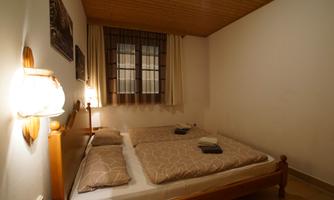 A cozy bedroom with a double bed and soft lighting. The walls are simple, and there is a window with curtains.