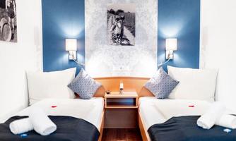 A modern room with two single beds, a bedside table, and stylish lamps. The walls are light blue with a decorative pattern and a piece of artwork.