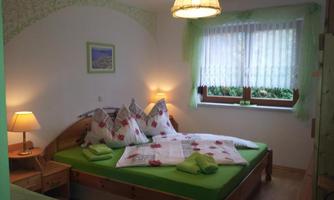 A cozy bedroom with a large double bed and green bedding. Pictures hang on the walls, and the window is decorated with curtains in a light green shade.
