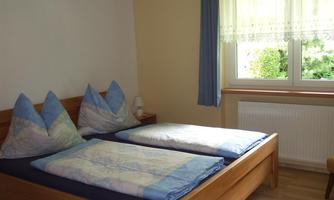 A simple bedroom with a double bed and two large pillows. Brightly lit by a window overlooking greenery.