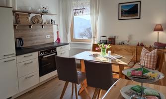 A modern kitchen with a dining table and chairs. Light comes through the window and there are flowers on the table.