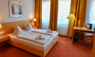 A cozy hotel room with two single beds and an orange carpet. The windows are decorated with light curtains, and there is a desk as well as lamps in the room.