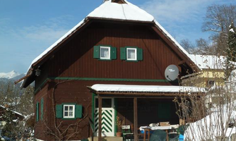 A cozy wooden house with a snow-covered roof and green shutters. The surroundings are wintery and idyllic.