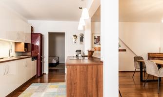 A modern kitchen with wooden and white furniture. The room is bright and inviting, with a cozy rug and a dining table set.
