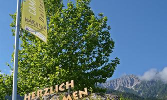 A welcome sign stands in front of colorful flowers and a large tree. In the background, mountains and a blue sky are visible.