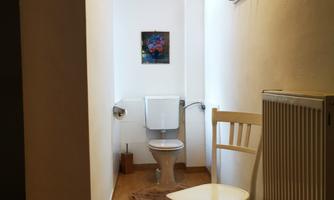A simple restroom with a white toilet and a chair. A small picture hangs on the wall, and a radiator is visible.