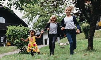 Three children run happily across a meadow in front of a house. They laugh and enjoy their time outdoors.