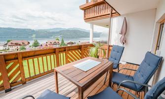 A cozy balcony with a wooden table and two chairs. In the background, a picturesque view of mountains and a body of water stretches out.