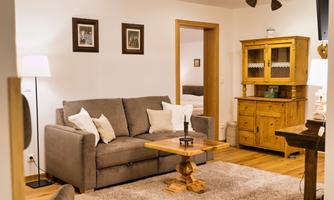 A cozy living room with a gray sofa and a wooden table. In the background, there is a sideboard and artistic wall paintings.