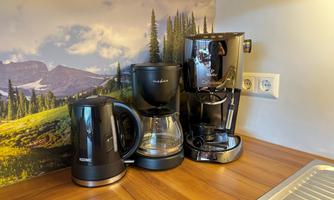 A modern kitchen unit with three coffee machines. In the background, a picturesque mountain landscape can be seen.