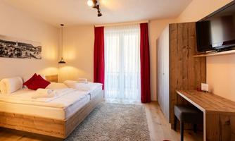 A cozy bedroom with two beds and wooden furniture. Large windows with red curtains provide plenty of daylight.