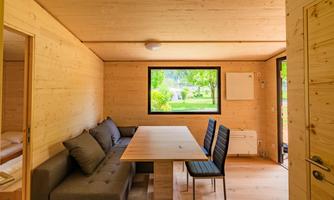 A modern room with wood paneling, a large table, and a cozy couch. Through the window, one can see a green landscape.