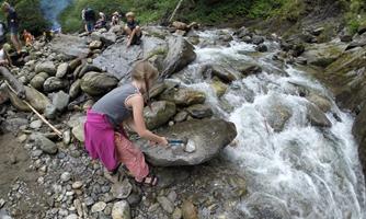 A child is sitting by a stream and exploring the stones. In the background, there are other people enjoying nature.