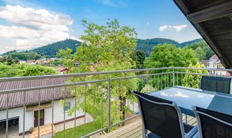 A terrace with a table and chairs offers a beautiful view of the green hills and trees. The sky is clear and slightly cloudy.