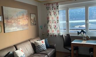 A cozy living room with a couch, a table, and large windows. There are decorative cushions and a painting on the wall.