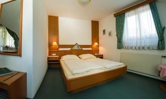 A simple hotel room with a double bed and wooden furniture. The room is bright and cheerfully furnished, with windows that let in daylight.