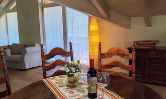 A cozy dining area with a round wooden table and four chairs. On the table, there is a bottle of wine and glasses, surrounded by warm light and a bright atmosphere.