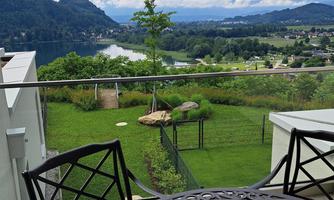 A balcony with a round table and chairs overlooks a green landscape and a river. In the background, mountains and a cloudy sky are visible.