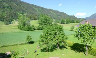 A picturesque landscape with green meadows and trees. In the background, gentle hills and wooded mountains can be seen.