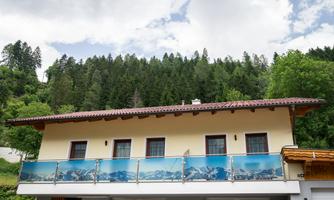 A modern house with a balcony that shows a view of the mountains. In the background, there are forested hills and a cloudy sky.