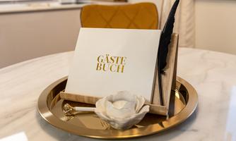An elegant guestbook on a golden tray, decorated with a white flower and a feather. In the background, a stylish chair is visible.