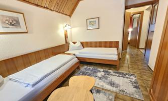 A simple room with two single beds and a small table. The room has wooden paneling and a cozy entrance.
