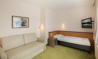 A simple hotel room with a single bed and a beige sofa. The walls are light and there are two pictures on the wall.