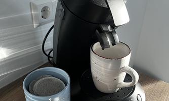 A coffee machine is sitting on a wooden board. Next to it are two cups and a wooden lid.