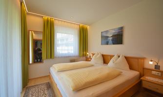 A cozy bedroom with a large bed and two white pillows. The windows are adorned with green curtains, and a mirror reflects the room.