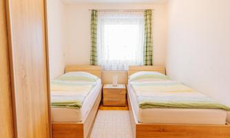 A simple bedroom with two single beds and a small nightstand. The room has a bright view through a window with curtains.