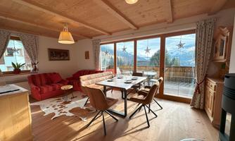 A cozy apartment with a wooden ceiling and large windows. The view shows snow-covered mountains and plenty of daylight.