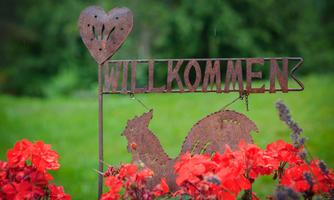 A welcoming metal sign with a heart and a chicken, surrounded by colorful flowers. It stands on a green lawn and conveys an inviting atmosphere.