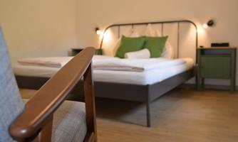 An inviting bedroom with a bed decorated with green pillows. Next to the bed is a nightstand, and in the foreground, there is a wooden chair.
