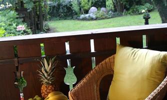 A cozy seating area on a balcony with a woven chair and colorful cushions. Next to the chair is a red table with fresh fruit and a view of a green garden.