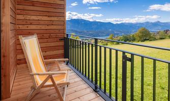 A cozy balcony with a wooden chair and a yellow backrest. In the background, green meadows and majestic mountains can be seen under a blue sky.