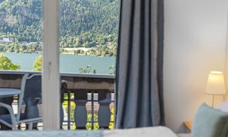 A view of a beautiful lake and the surrounding forests from a room. The windows overlook a cozy balcony with chairs.