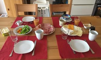 A set dining table with several plates and glasses. There are sausages, cheese, vegetables, fruit, and drinks.