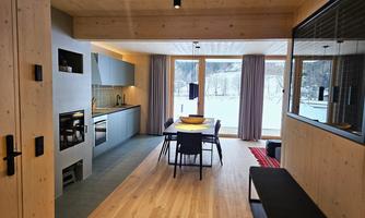 A modern kitchen with a dining area and large windows. The view shows the snow and nature outside.