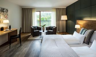 A modern hotel room with a large bed and cozy seating areas. The room has large windows overlooking nature and is tastefully decorated.