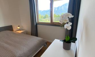 A bright bedroom with a bed and a nightstand. Through the window, you can see a beautiful mountain landscape.