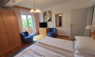 A cozy hotel room with two beds, a TV, and two blue chairs. The balcony door lets in plenty of daylight and offers a view of the greenery.