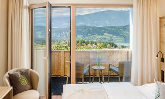 A cozy room with a view of the mountains and a balcony. The view shows a green landscape and a clear sky.